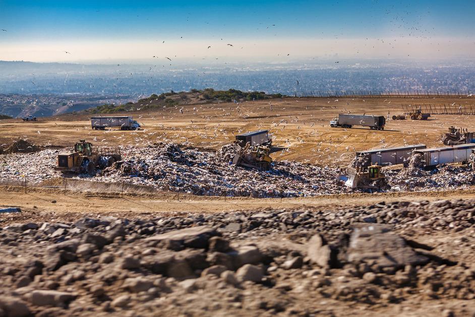 Trucks surround a landfill on the top of a sandy mountain while birds fly in the air.
