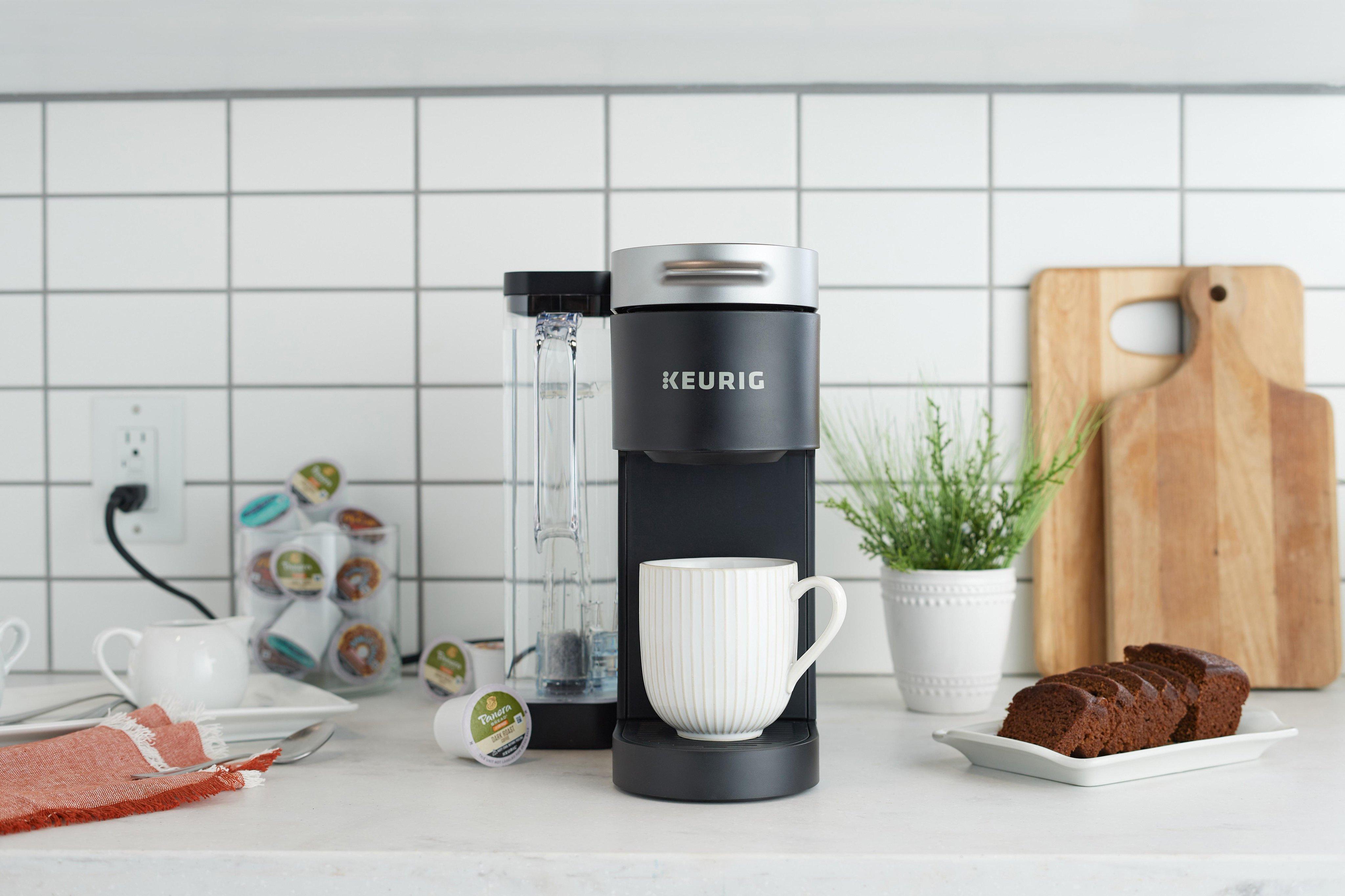 A Keurig machine is pictured atop a kitchen counter beside a chocolate pastry, cutting board, K-Cups, and other accessories.