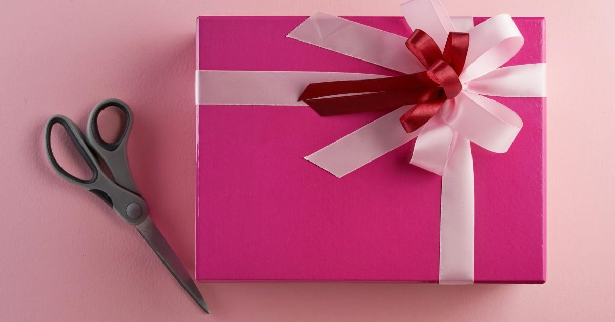 A pink box tied with white and red ribbons sits on a table next to a pair of scissors